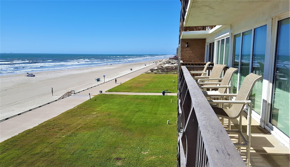 A spacious wrap-around patio offers panoramic views of the Gulf Coast, with multiple Captain chairs facing the ocean. The patio seamlessly connects to well-maintained grassy areas and a pathway leading to the beach. The coastline features gentle waves and a sandy expanse under a clear blue sky.