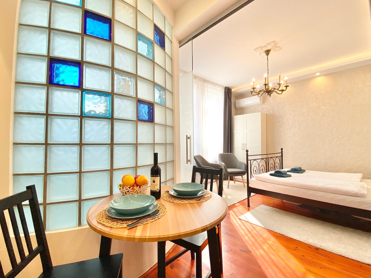 A bright studio apartment features a dining table set for two, including plates and a fruit basket. The living area includes a spacious bed and two comfortable armchairs. A decorative glass wall allows light to filter through, contributing to the open atmosphere.