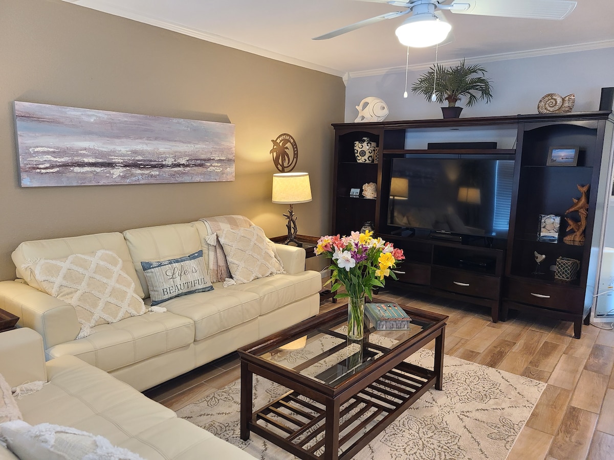 A light-colored living area is featured, showcasing a seating arrangement with a cream-colored sofa and a dark wooden coffee table. A flat-screen television is positioned on a dark wooden console. A floral centerpiece adds a touch of color, while warm lighting creates a cozy environment.
