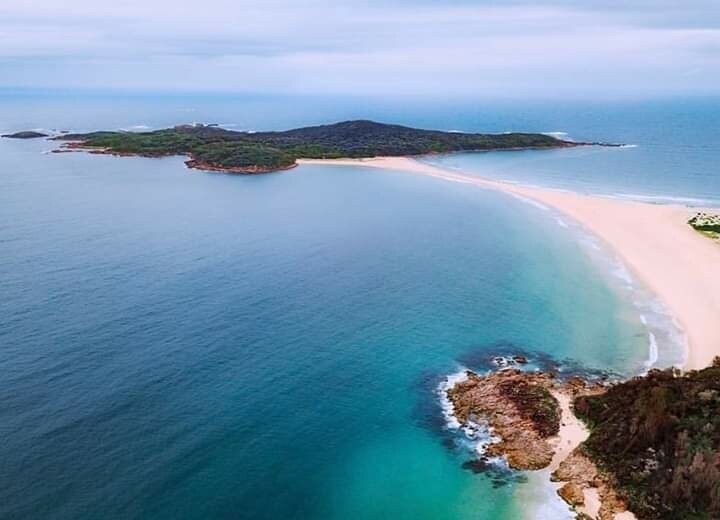 An aerial view captures a serene coastal landscape, featuring a sandy beach curving into the turquoise waters. Lush green hills are visible in the distance, contrasting with the rocky shoreline. Gentle waves lap against the beach, enhancing the tranquil atmosphere of the scene.