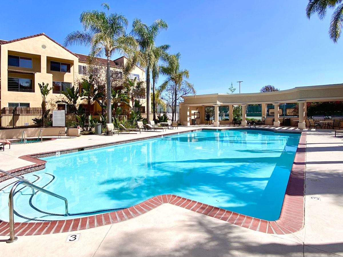 A gated swimming pool is surrounded by palm trees and lounge chairs. The clear blue water reflects the sunlight under a bright sky, inviting relaxation in the spacious outdoor area. A shaded seating area and a terrace are visible in the background.