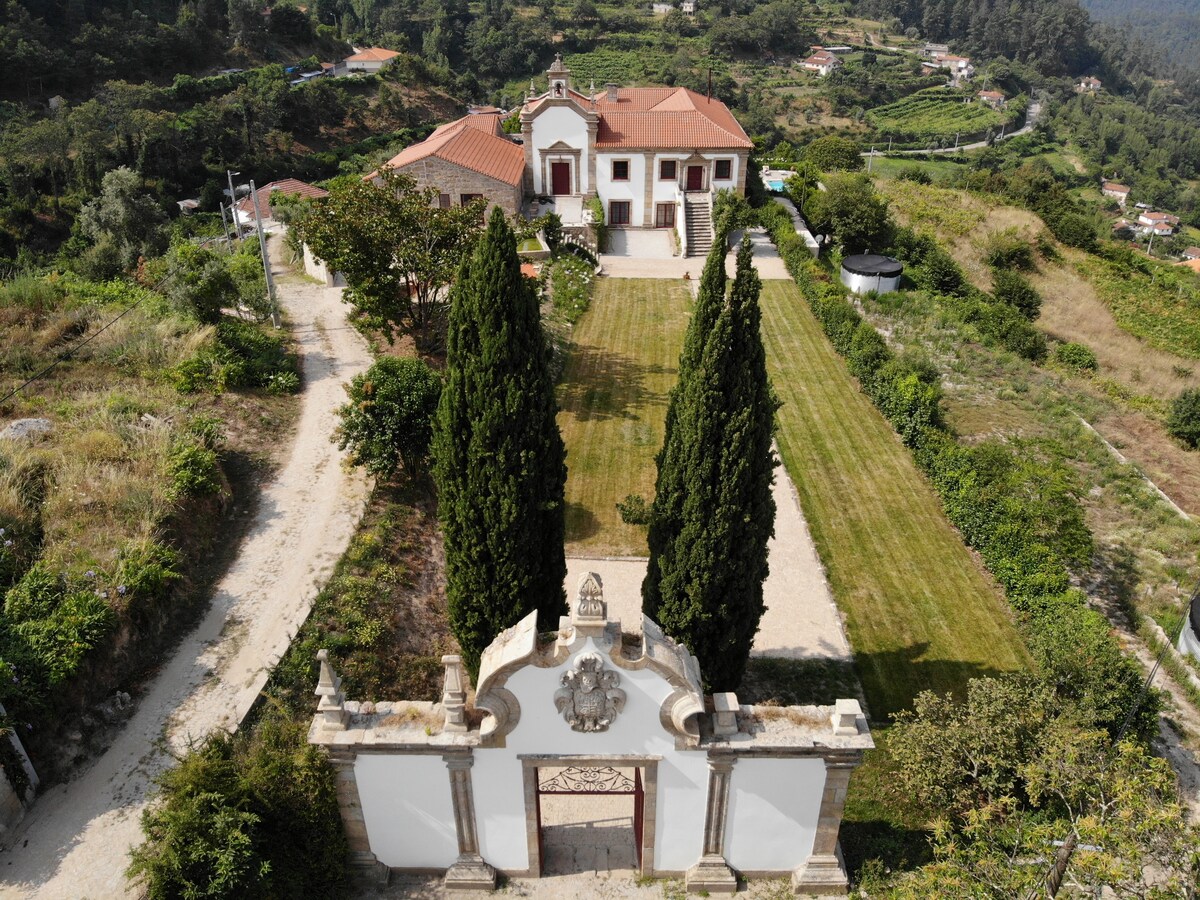 An aerial view showcases a historic 17th-century solar surrounded by lush greenery and rolling hills. The manicured lawn is framed by tall cypress trees, leading to a grand entrance with ornate stonework. The charming building features a red-tiled roof and multiple windows, blending harmoniously with the landscape.
