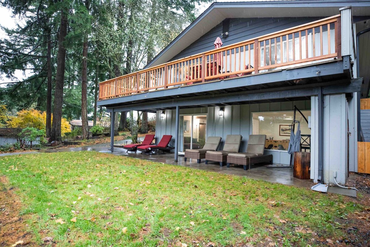 The exterior view displays a modern two-story home with a spacious upper deck and patio seating. A lush green lawn surrounds the lower patio, accentuated by comfortable lounge chairs. The entrance features large windows, allowing natural light to enter the house.