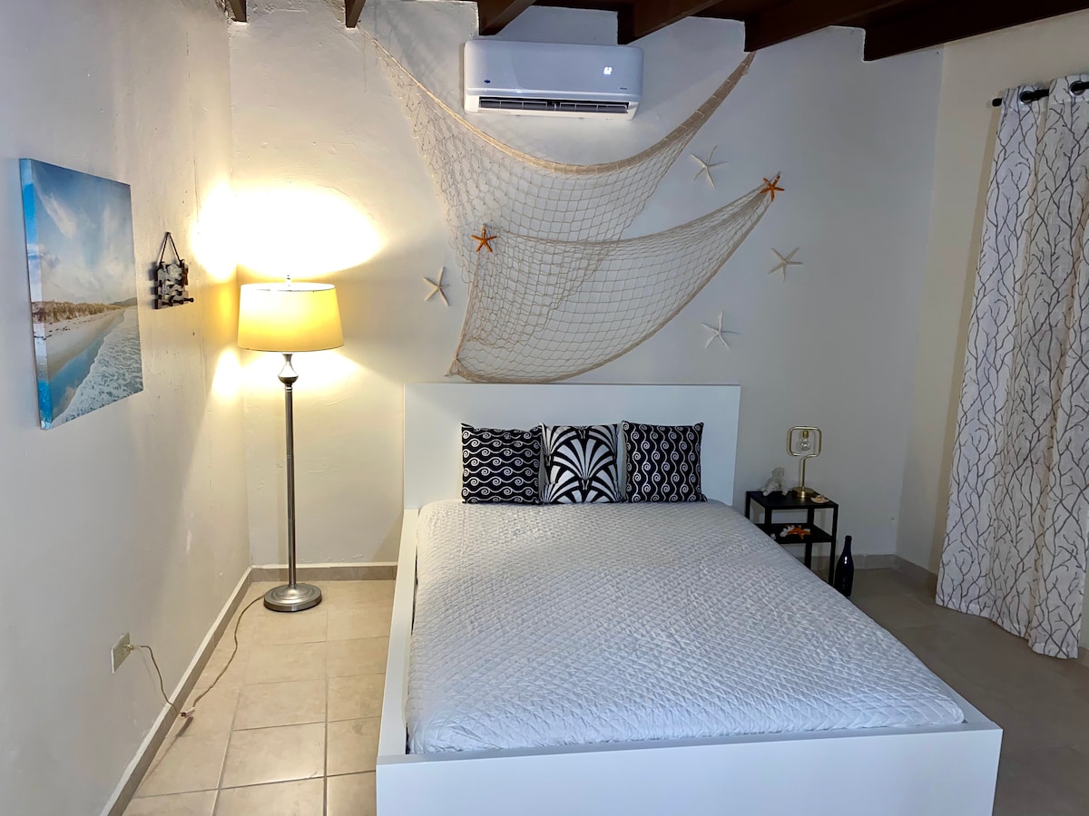 A bright bedroom features a spacious bed with decorative pillows against a simple backdrop. A lamp provides soft lighting, while a wall adorned with a fishing net and starfish adds a coastal touch. A window is covered with patterned curtains, allowing natural light to enter.