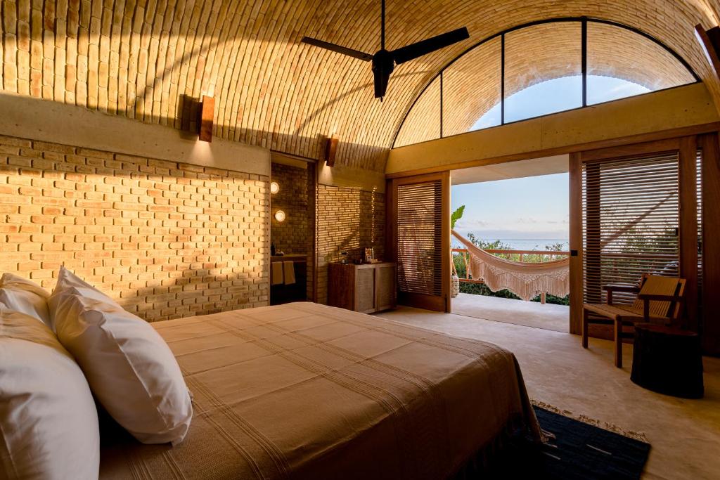A spacious bedroom features a vaulted ceiling with natural brick walls. A king-size bed is centered in the room, while a large glass door opens to a terrace with a hammock, allowing natural light to fill the space.