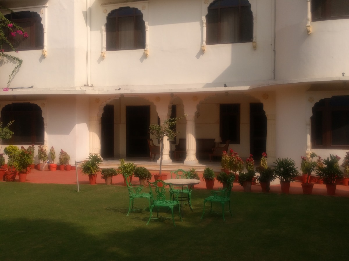A well-kept garden area is featured, with green seating arranged around a small table. The outdoor space is surrounded by colorful potted plants and provides access to the bungalow's white facade with arched windows.