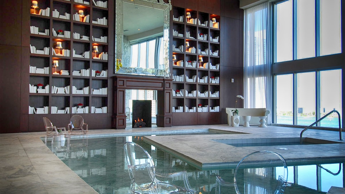 A spacious spa area features a calming indoor pool with clear water. Stylish lounge chairs are placed on a marble surface beside the pool. A large mirror and bookshelves line one wall, while floor-to-ceiling windows allow natural light to fill the space.