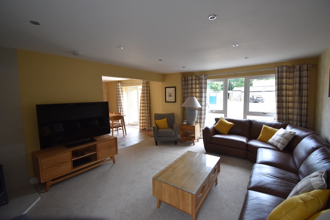 A spacious living area is furnished with a large brown corner sofa and an elegant wooden coffee table. A wide-screen TV is mounted on the wall, and light streams through windows dressed in plaid curtains, illuminating the cozy space.