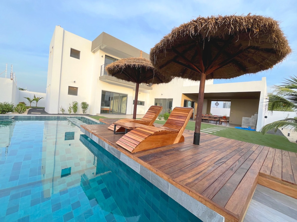 A private pool area features two wooden lounge chairs under thatched umbrellas, surrounded by a wooden deck. The pool's clear water reflects the sky, while the modern villa stands in the background, offering a blend of indoor and outdoor living spaces.