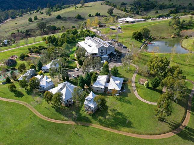 An aerial view of the resort showcases landscaped grounds with walking paths winding through green lawns. Various chalet-style accommodations are surrounded by trees, and a pond reflects the surrounding nature. The main building is visible in the background, providing access to amenities.