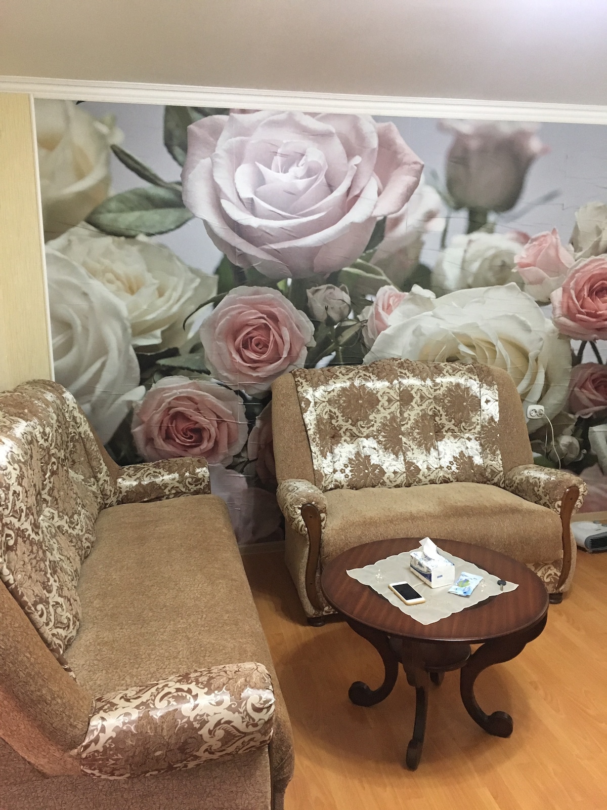 A comfortable seating area is shown, featuring a pair of upholstered sofas in a light brown tone, complemented by a round wooden coffee table. The wall behind is adorned with a floral mural showcasing soft pink and white roses, creating a serene backdrop.
