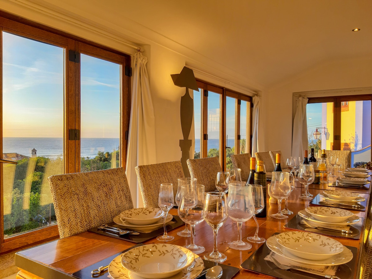 A dining area features a long wooden table set with plates, glasses, and cutlery, overlooking expansive ocean views through large glass windows. Natural light fills the space, which is enhanced by soft fabrics and elegant decor.