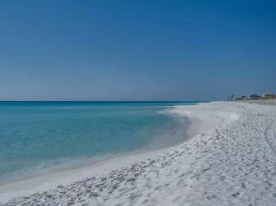 A serene beach scene showcases expansive stretches of sugar-white sand meeting the clear turquoise waters of the Gulf of Mexico. The horizon is marked by a gentle gradient of blue, with a few distant structures subtly visible along the shore.
