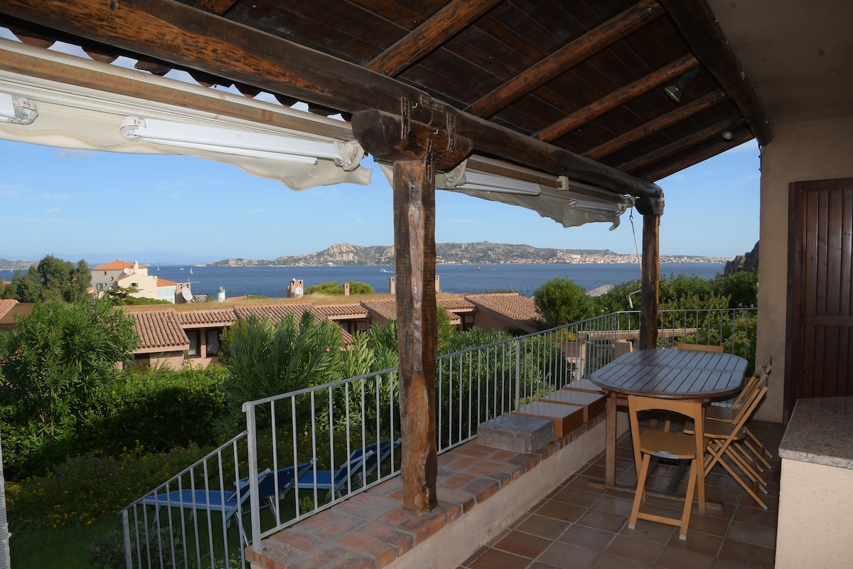 A covered terrace features a wooden table and chairs, offering a view of the sea and surrounding landscape. The terrace is enclosed by stone walls and wooden posts, with greenery visible in the foreground and distant coastal hills adding to the scene.