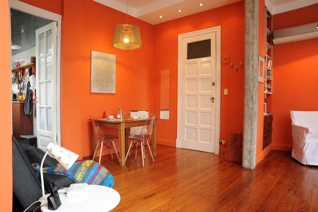 Sofá cama, vista a la cocina y puerta de entrada. Mesa con lámpara y sillas de estilo escandinavo.