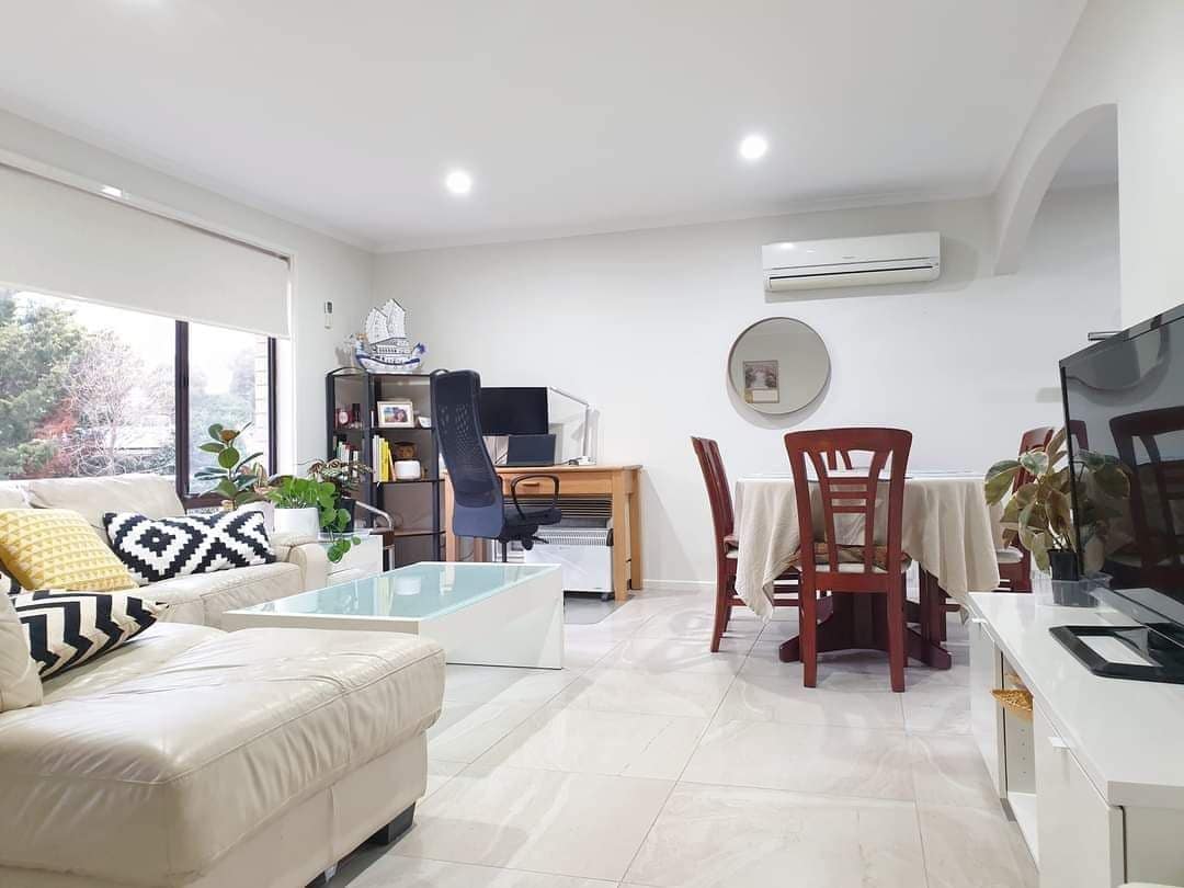 A modern living area is shown with a comfortable light-colored sofa and a low glass coffee table. A dining table surrounded by wooden chairs is visible in the center. A work desk with a chair and decorative plants add to the inviting ambiance.