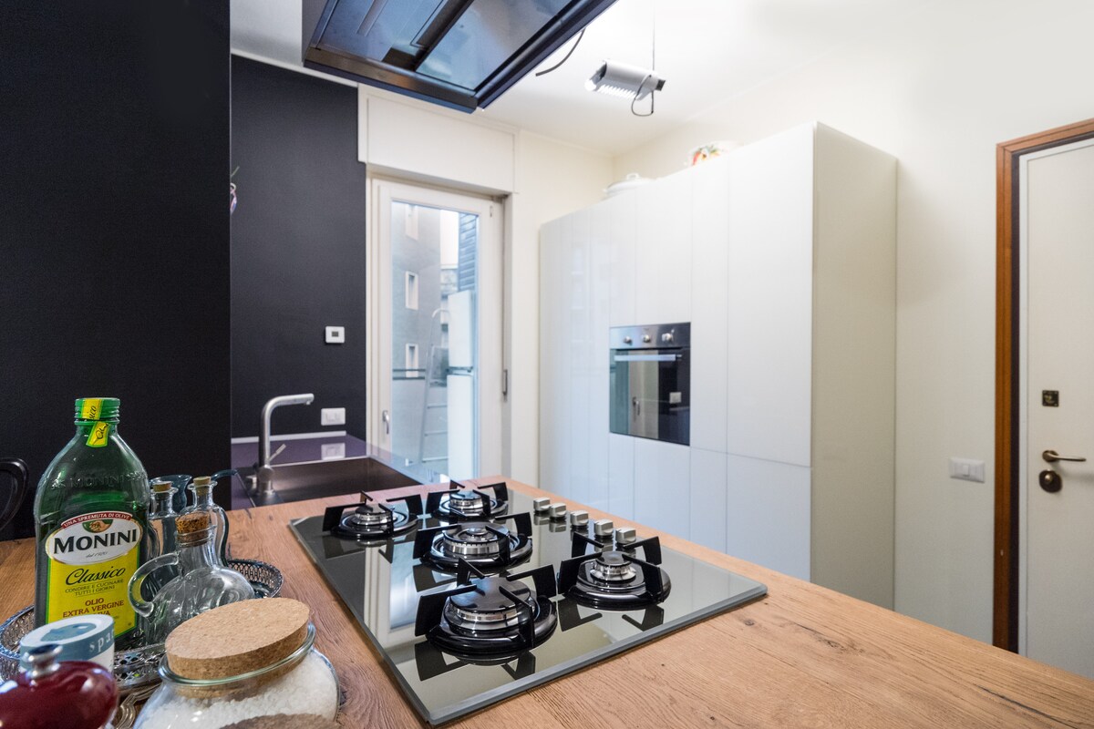 A modern kitchen is showcased, featuring a sleek design with white cabinetry and a spacious wooden countertop. A gas cooktop is prominently displayed, surrounded by cooking essentials. Natural light enters through a window, illuminating the organized space.