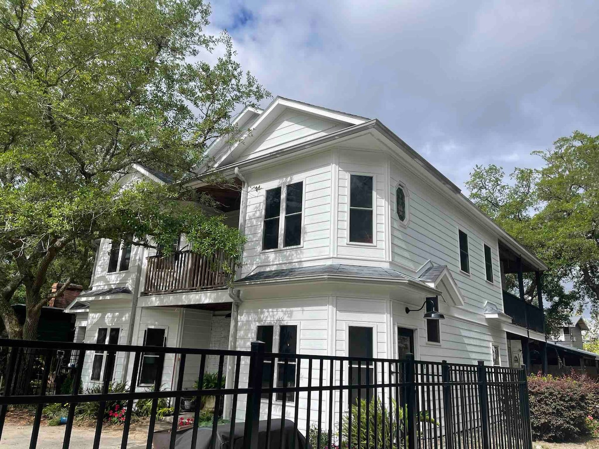The exterior of a two-story white house is displayed, framed by lush greenery. A covered balcony is located on the upper level, while dark windows reflect the surroundings. A black fence lines the property, adding a sense of privacy and security.