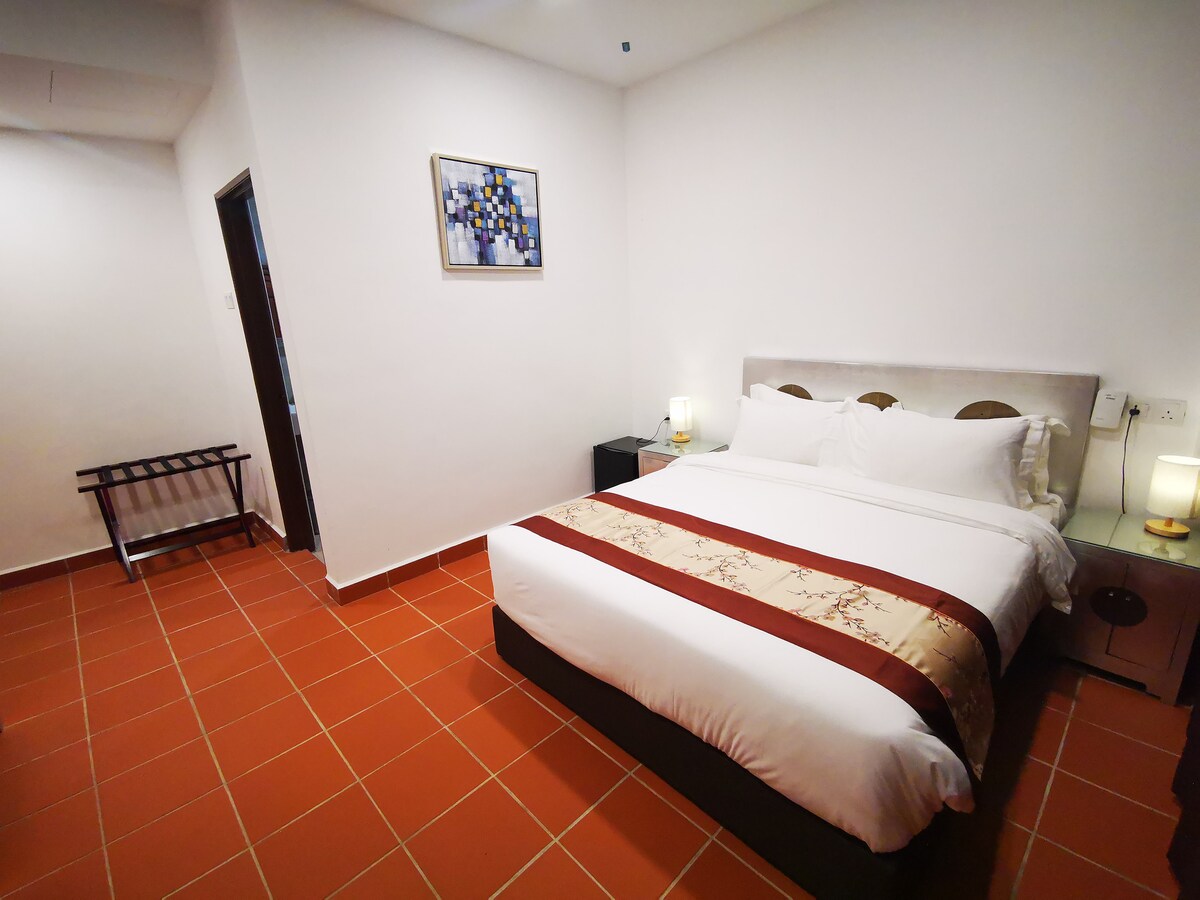 A spacious sleeping area is presented, featuring a comfortable bed with crisp white linens and a decorative brown and gold bed runner. Soft lighting is provided by two bedside lamps, and a piece of wall art adds a touch of color. The tiled floors enhance the welcoming ambiance.