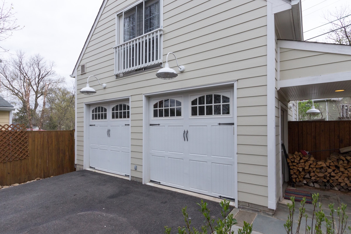 The exterior view features a light-colored garage with two large, white double doors. Decorative windows are positioned above the doors, alongside wall-mounted lighting fixtures. A neatly paved driveway is shown, and a wooden fence can be seen in the background, enhancing the property’s enclosed feel.