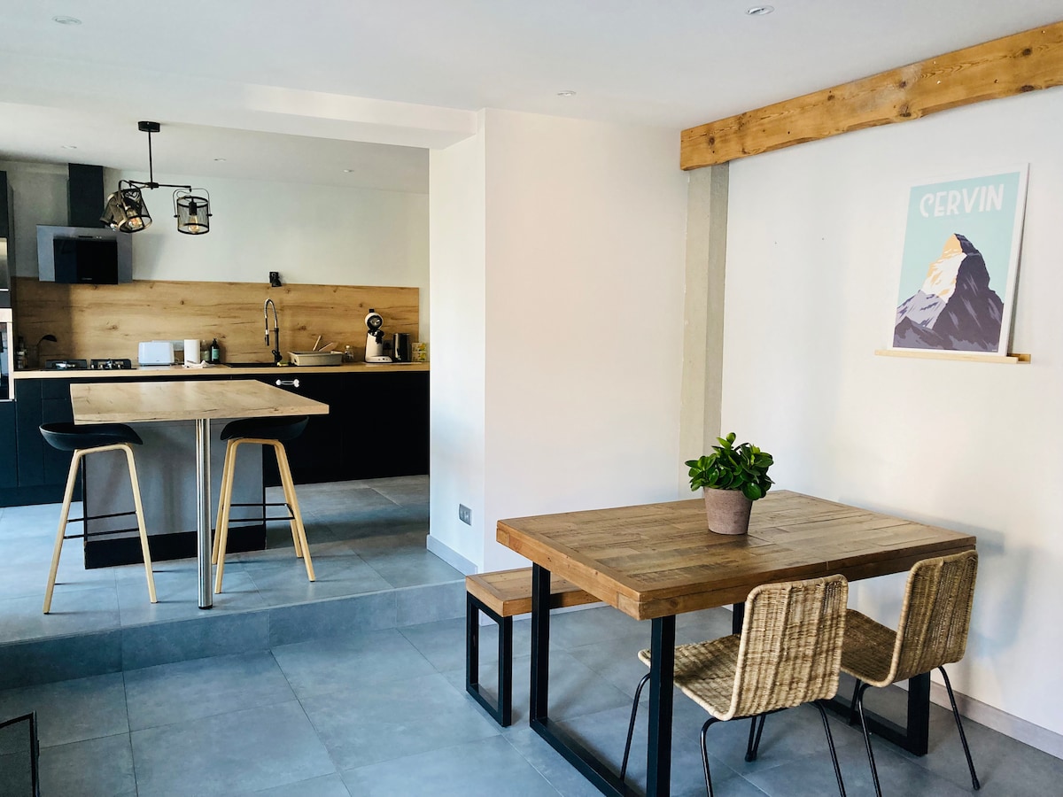 A modern kitchen and dining area are showcased, featuring a black cabinetry and wooden countertops. A dining table with a potted plant is visible, accompanied by two woven chairs. The kitchen area is equipped with essential appliances and illuminated by pendant lighting.