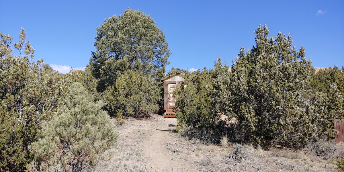 A rustic structure is surrounded by a mix of Junipers and Pinion pines, with a clear pathway leading to the entrance. The blue sky contrasts against the greenery, creating a serene outdoor setting.