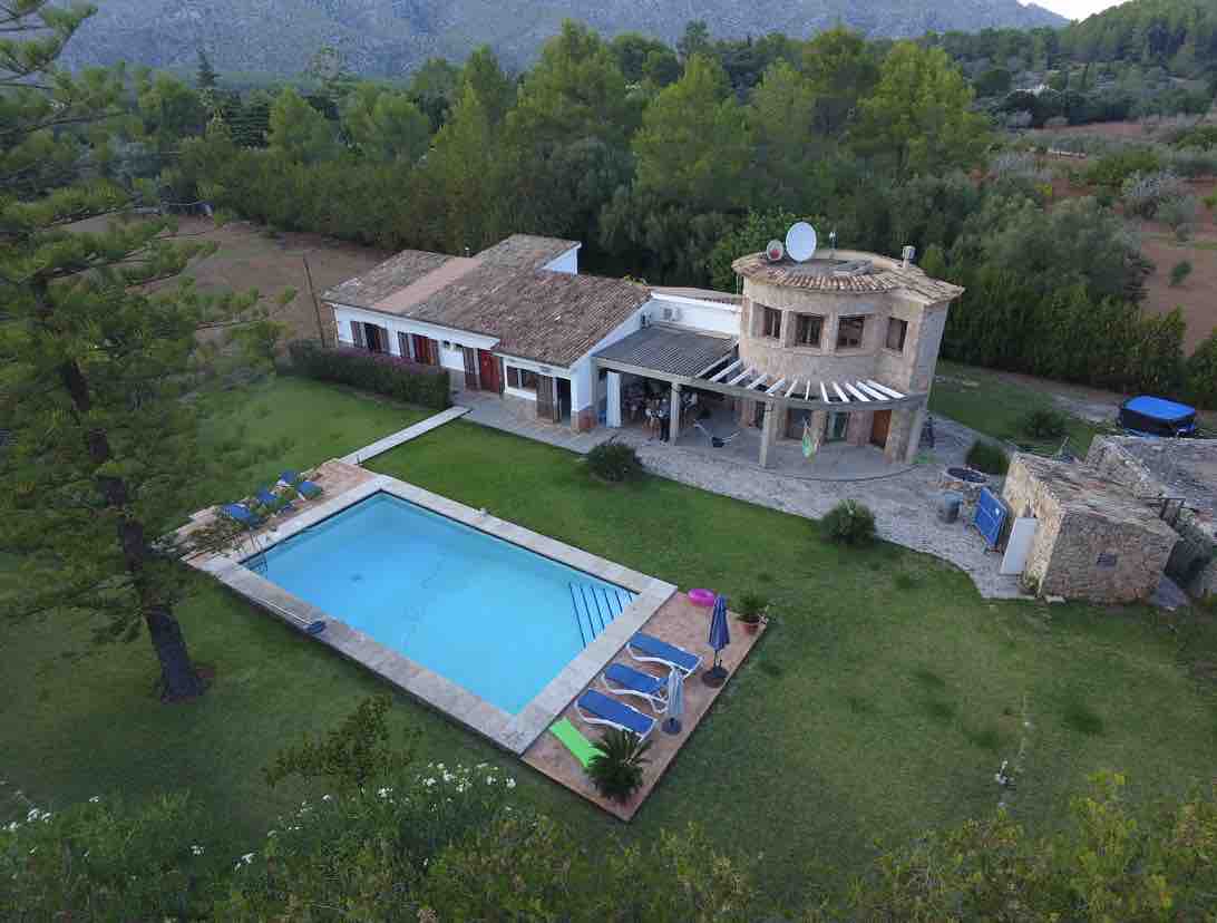 An aerial view of the villa showcases a spacious garden and a large, inviting swimming pool. Surrounding trees provide shade, while lounge chairs are arranged thoughtfully around the pool area. The two-story villa features a traditional stone exterior and multiple outdoor seating areas under awnings.