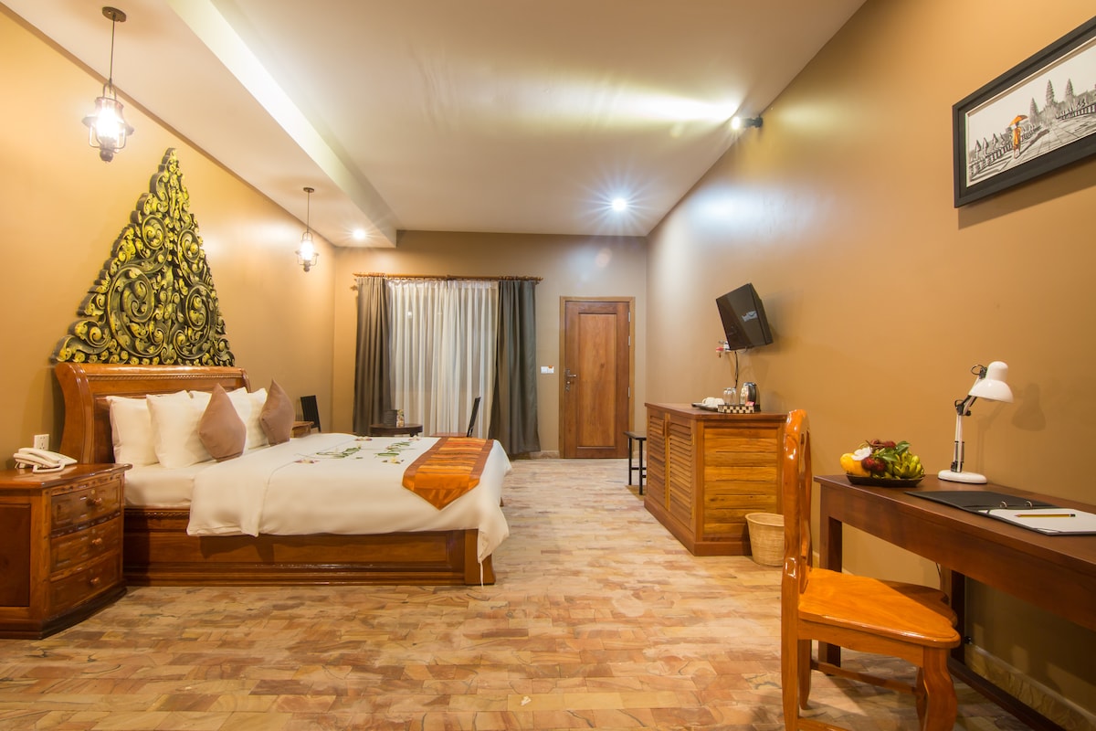 A spacious room features a wooden bed adorned with decorative accents and fresh flowers. A study desk with a lamp and a wooden chair sit in one corner, while a welcoming arrangement of fruit is displayed nearby. Soft lighting and warm-colored walls create a calming ambiance.