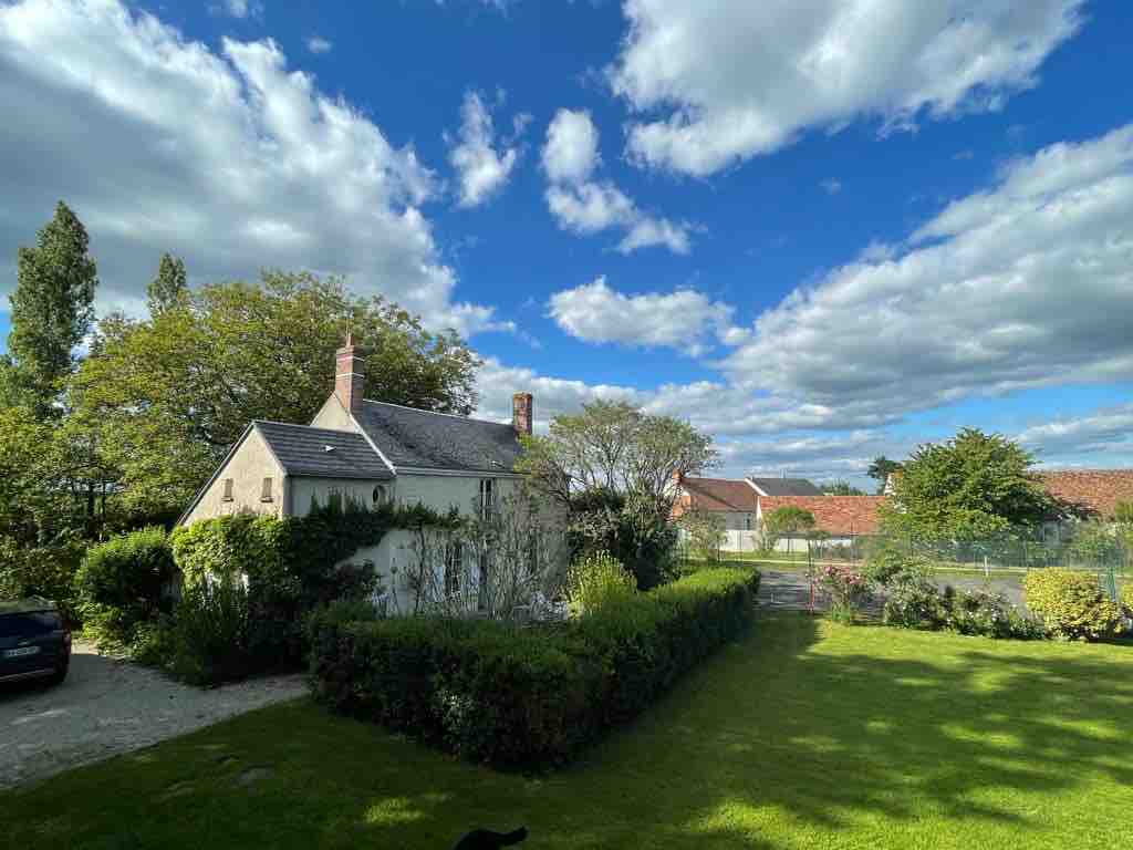 A charming house is surrounded by a lush garden and vibrant greenery. The sky is filled with fluffy clouds, while the sunlight casts a warm glow on the well-maintained landscape, creating a serene outdoor atmosphere.