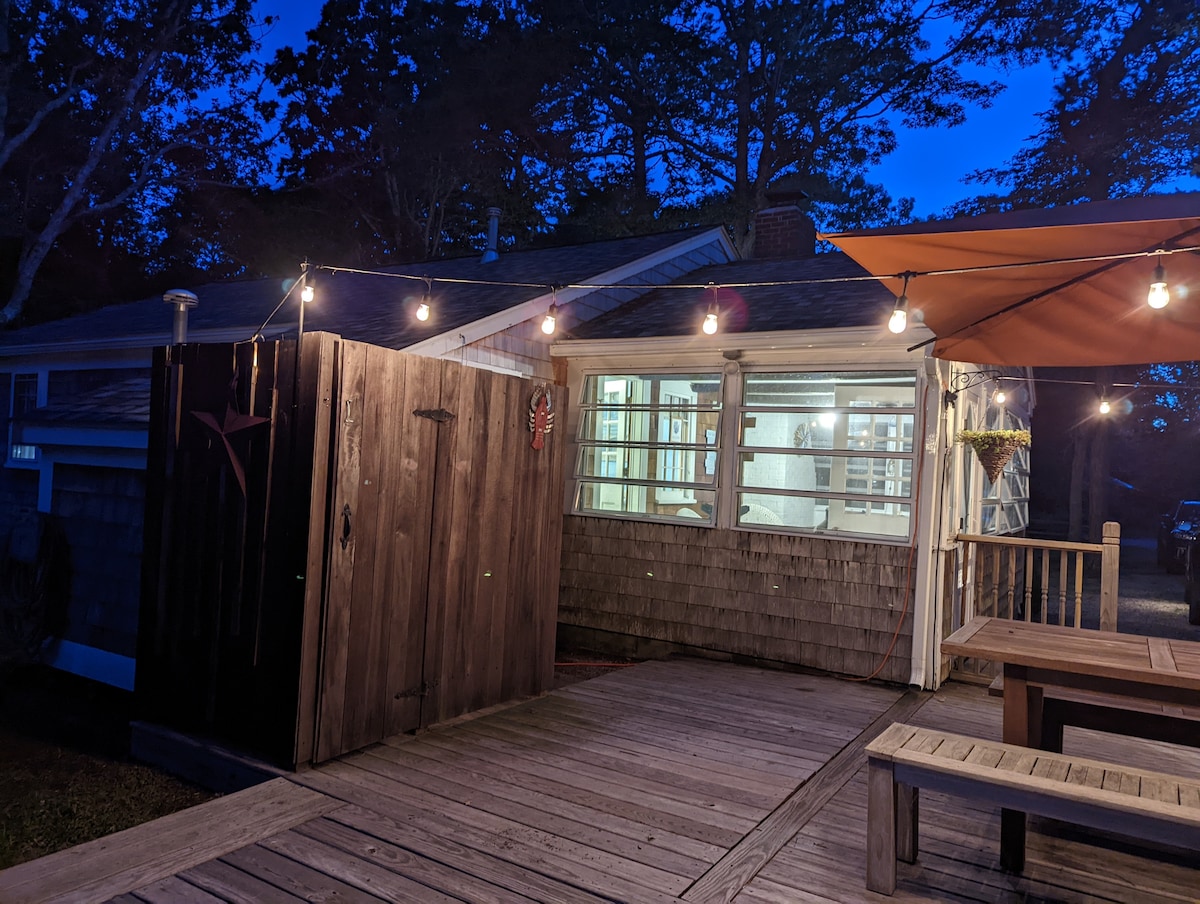 An outdoor deck is illuminated by warm string lights, highlighting a private outdoor shower area with wooden paneling. The adjoining enclosed porch features large windows that allow visibility into the cottage's living space, presenting a cozy and inviting entrance.