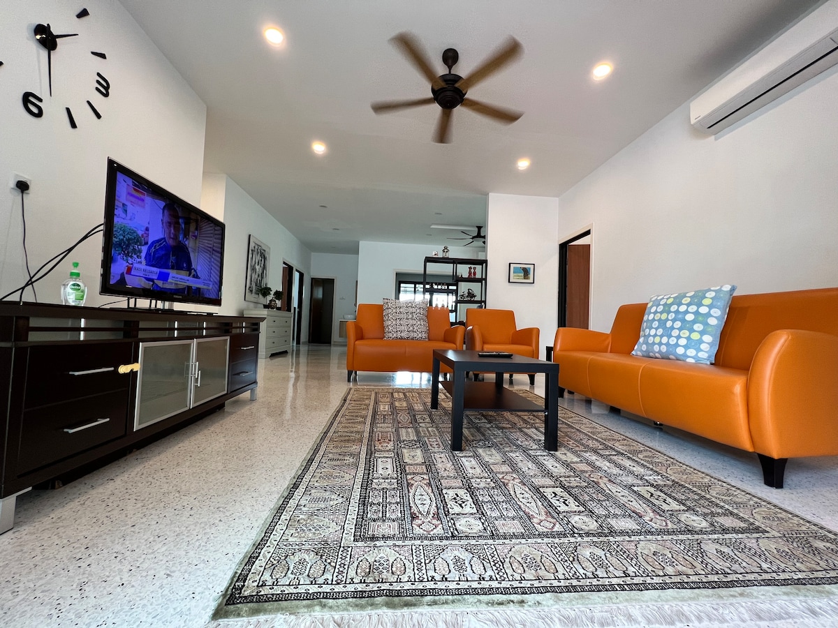 A spacious living area is highlighted by bold orange sofas and a coffee table set on a textured area rug. A sleek television unit holds a flat-screen TV, while a ceiling fan offers comfort. Natural light fills the room from unseen windows.