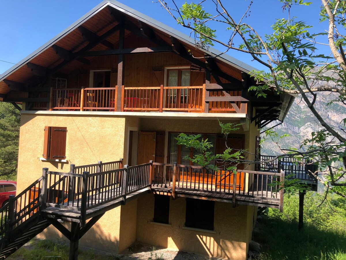 A wooden chalet is situated in a natural setting, showcasing multiple balconies with wooden railings. The exterior features a combination of warm earthy tones and natural wood, surrounded by greenery. Mountain views are visible in the background, contributing to the serene environment.