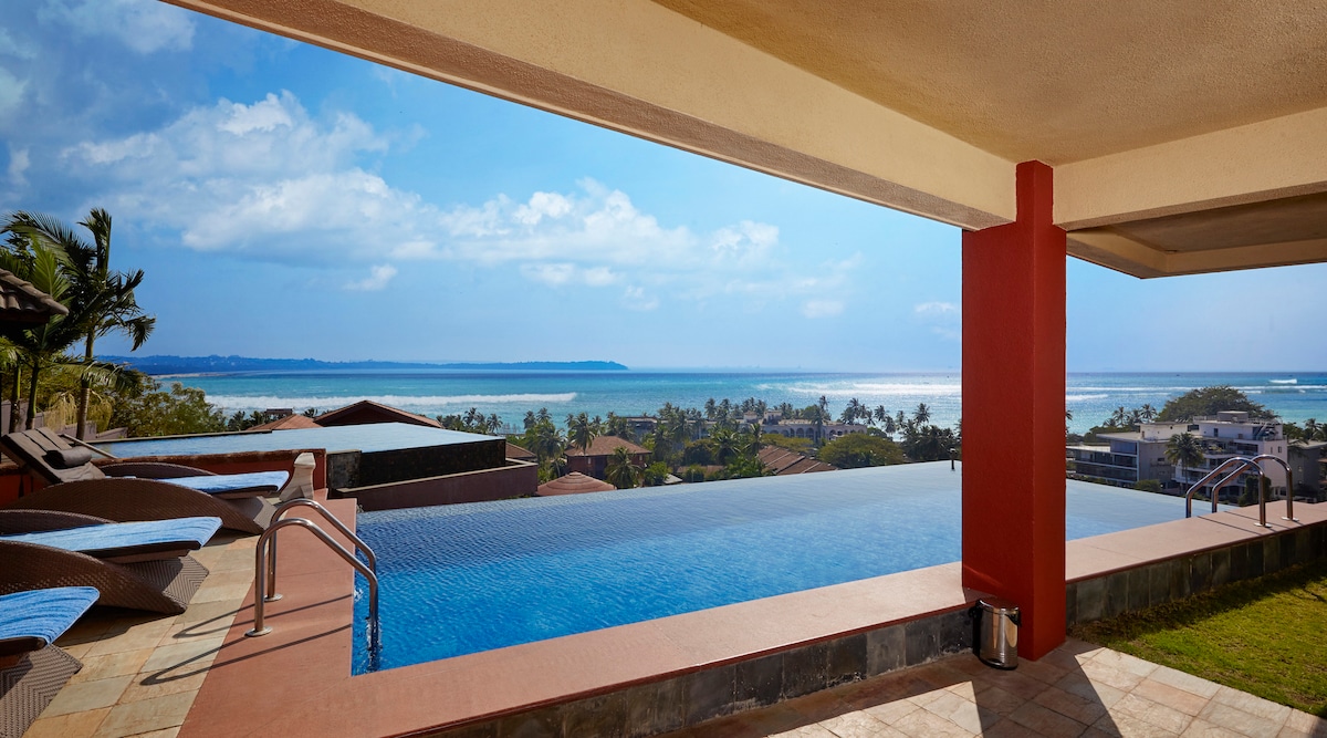 An infinity pool extends towards a breathtaking view of the Arabian Sea, surrounded by lush greenery and distant hills. The tranquil blue waters seamlessly blend with the ocean horizon, while a shaded overhang provides a cooling space nearby, enhancing the relaxed atmosphere of the outdoor area.