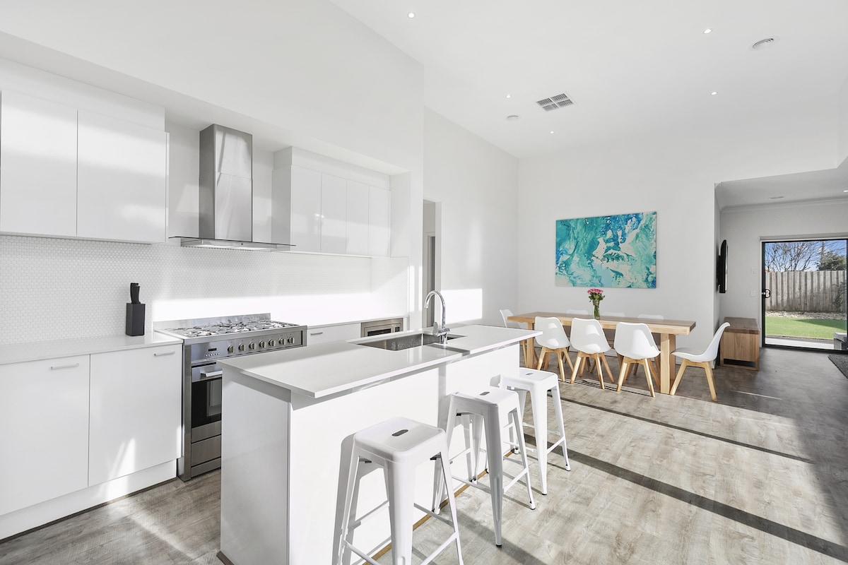A modern kitchen is highlighted by sleek white cabinetry and stainless-steel appliances. A large island with seating is positioned at the center. Natural light floods the space, illuminating a dining area visible in the background, featuring a wooden table and white chairs.
