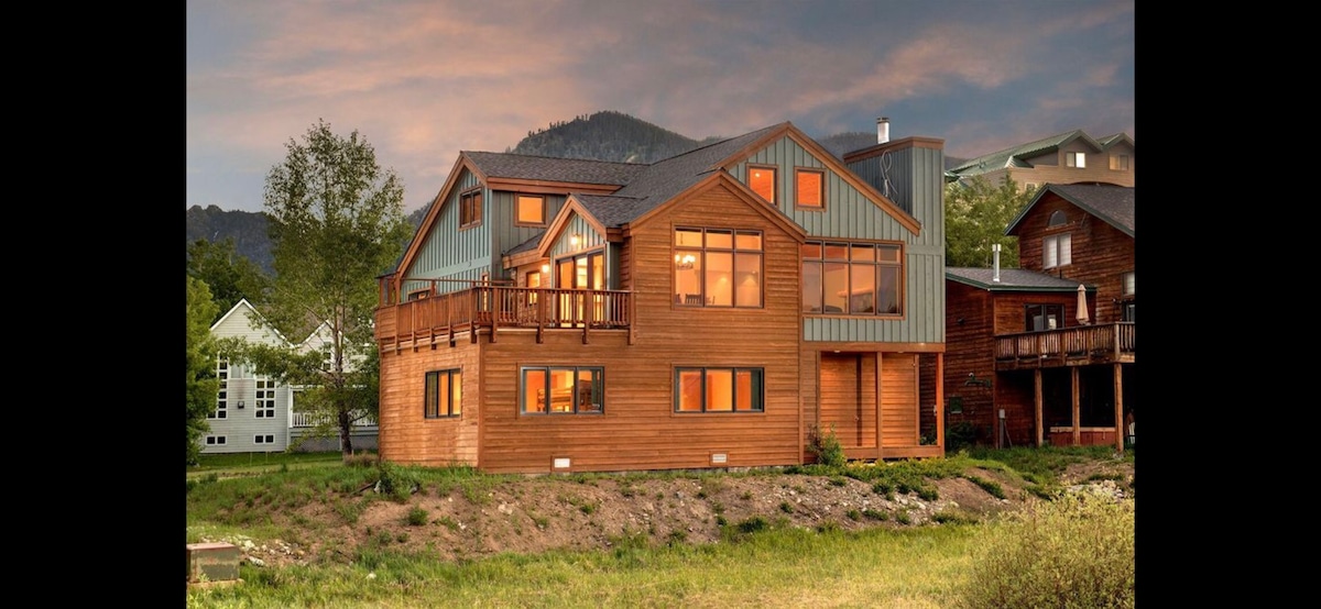 The lakeside retreat features a beautiful wooden exterior with large windows reflecting the soft hues of the sunset. A spacious deck extends from the building, providing outdoor space with scenic views of the surrounding landscape and mountains.