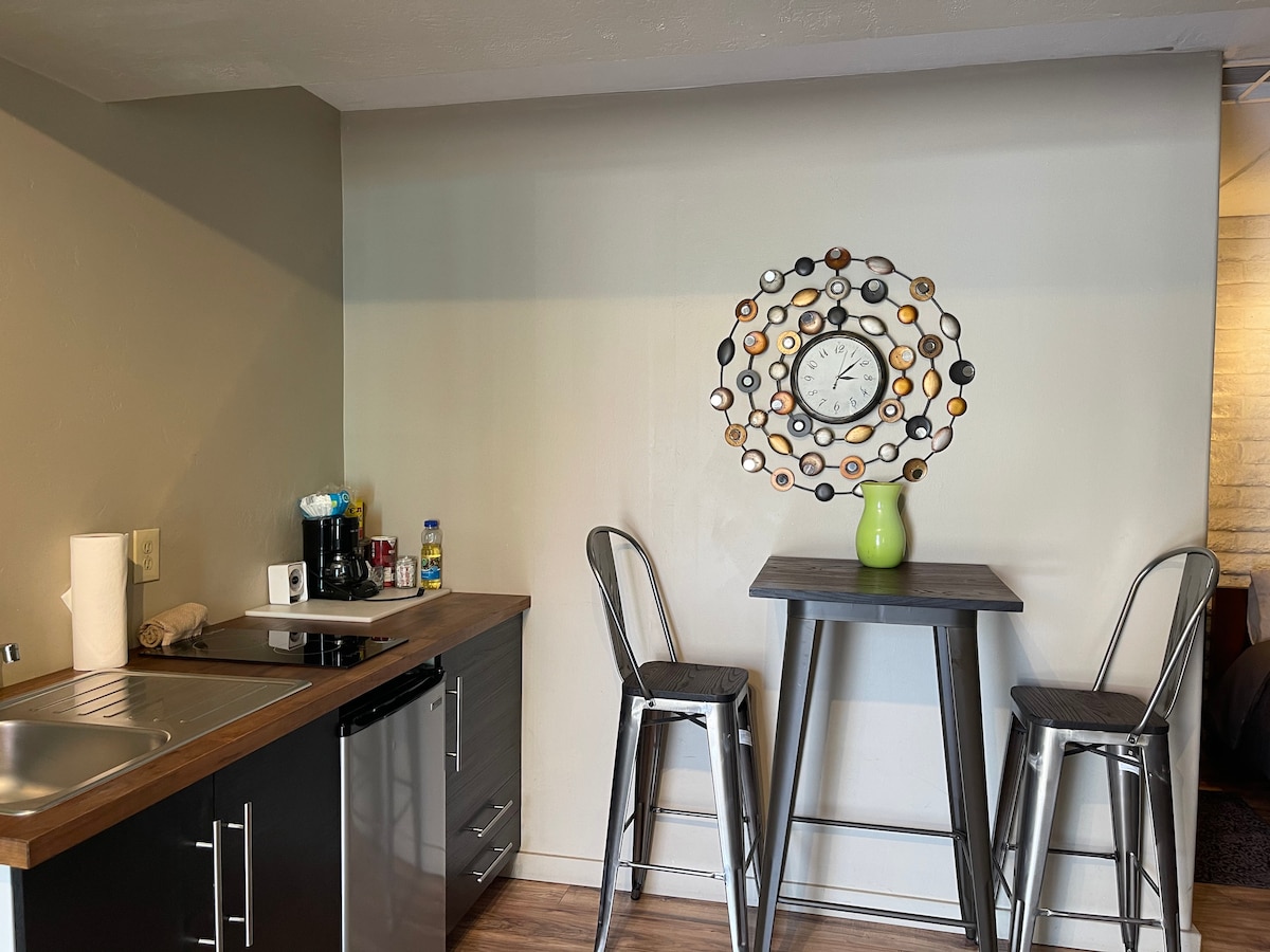 A compact kitchen area features a modern stainless steel sink and mini refrigerator. A wooden countertop is paired with two tall metal stools at a small table. A decorative wall clock is displayed above a green vase, adding a touch of color to the neutral walls.