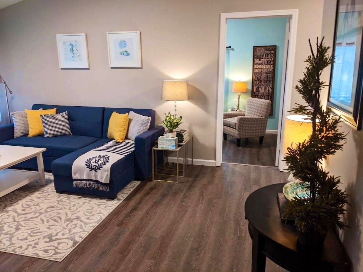 A cozy living space features a blue sectional sofa adorned with yellow accent pillows. A decorative table and lamp provide soft lighting, while a patterned area rug adds warmth. A framed artwork adorns the wall, and an entrance leads to a small seating area.