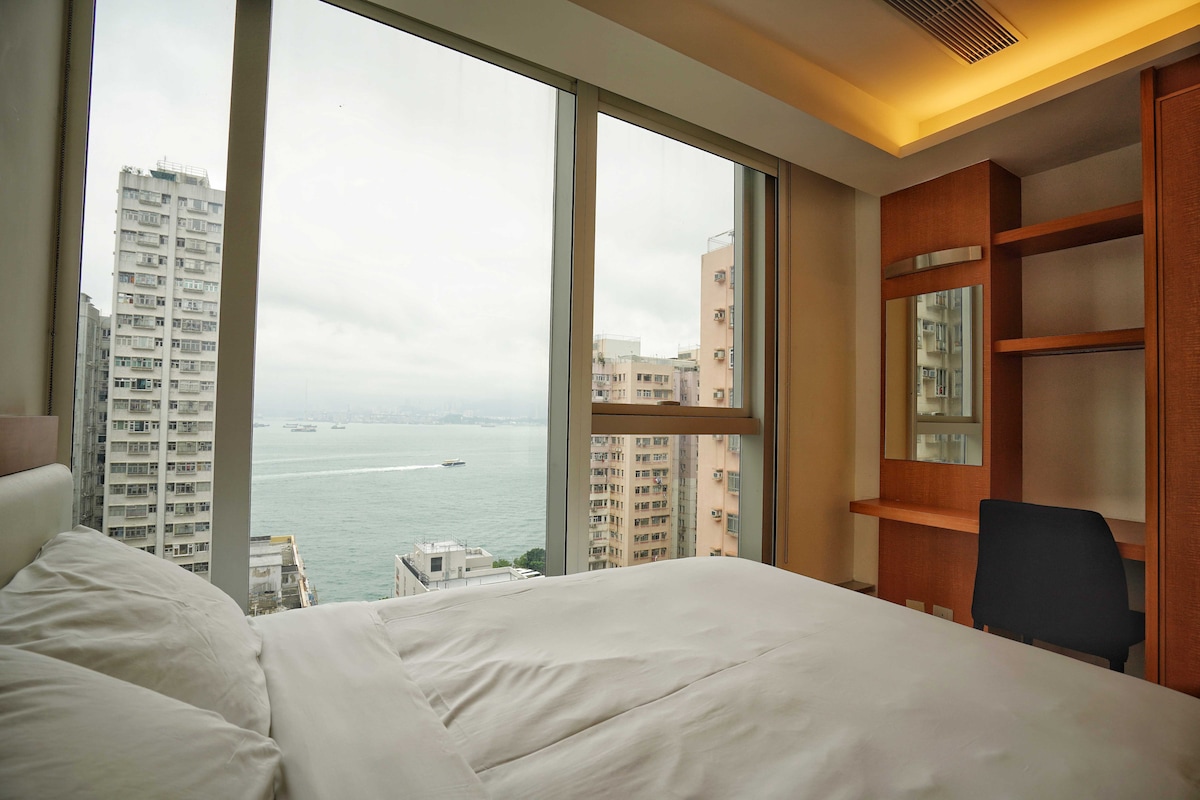 A serene bedroom features a spacious bed with crisp white bedding, alongside a wooden desk and chair. Large windows provide a panoramic view of the harbor, with buildings visible in the foreground and a cloudy sky above.