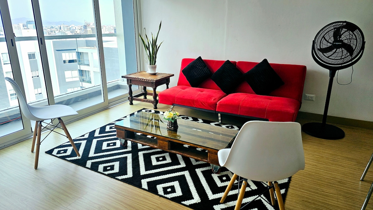 A bright living area features a red sofa adorned with black cushions, accompanied by two white chairs. A low wooden coffee table sits on a geometric patterned rug. Large glass doors offer views of the surrounding cityscape, complemented by a green plant and a standing fan.