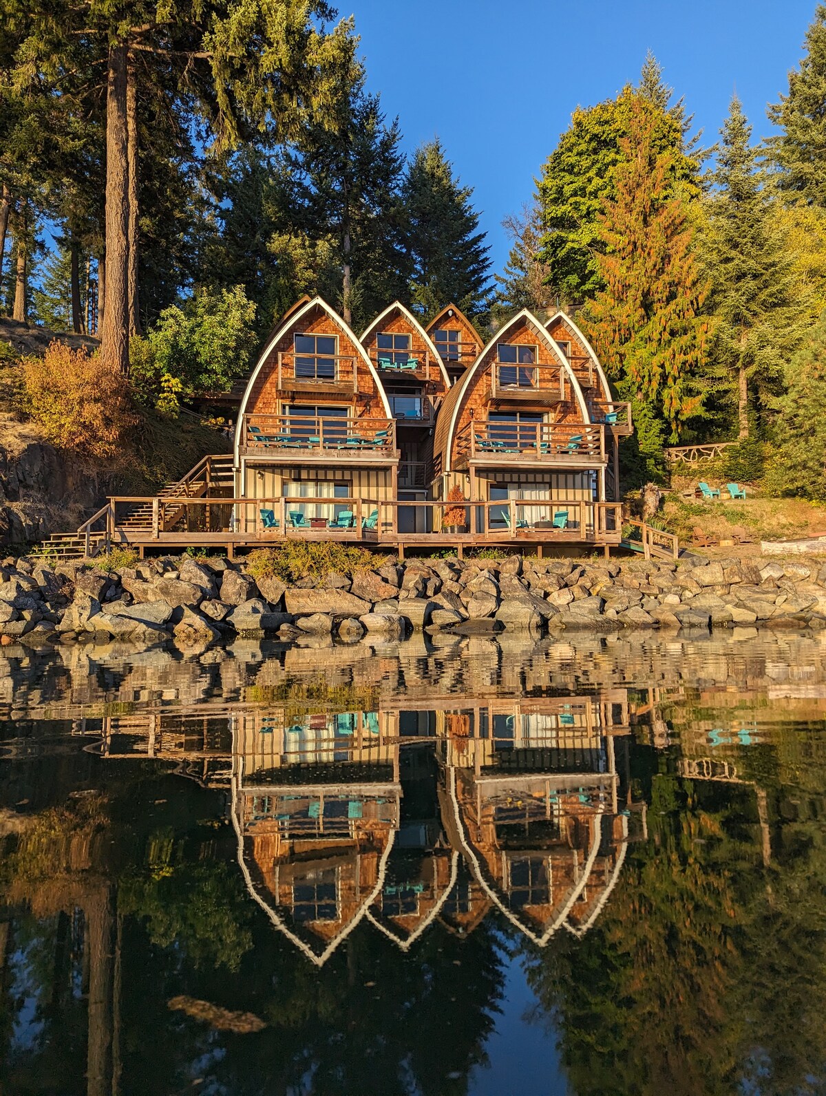 The exterior of a distinctive two-story A-frame chalet is reflected in calm waters. The building features a unique curved roof resembling a ship's bow, surrounded by tall trees. A wooden deck with seating provides outdoor space, enhancing the natural setting.