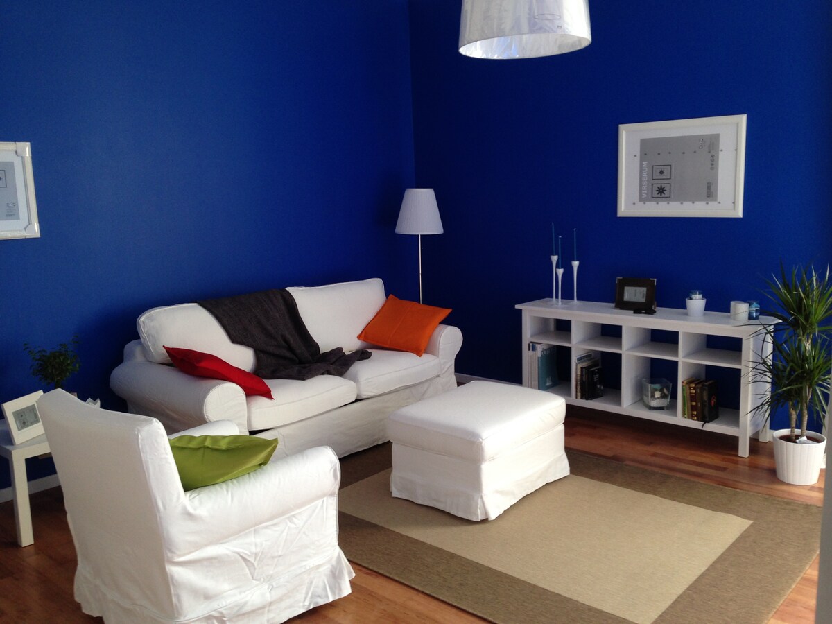 A bright living space features bold blue walls complemented by a crisp white sofa and chair. An orange and green pillow add subtle color, while a white coffee table sits center stage. A modern lamp and a minimalist shelf with decorative items contribute to the inviting atmosphere.