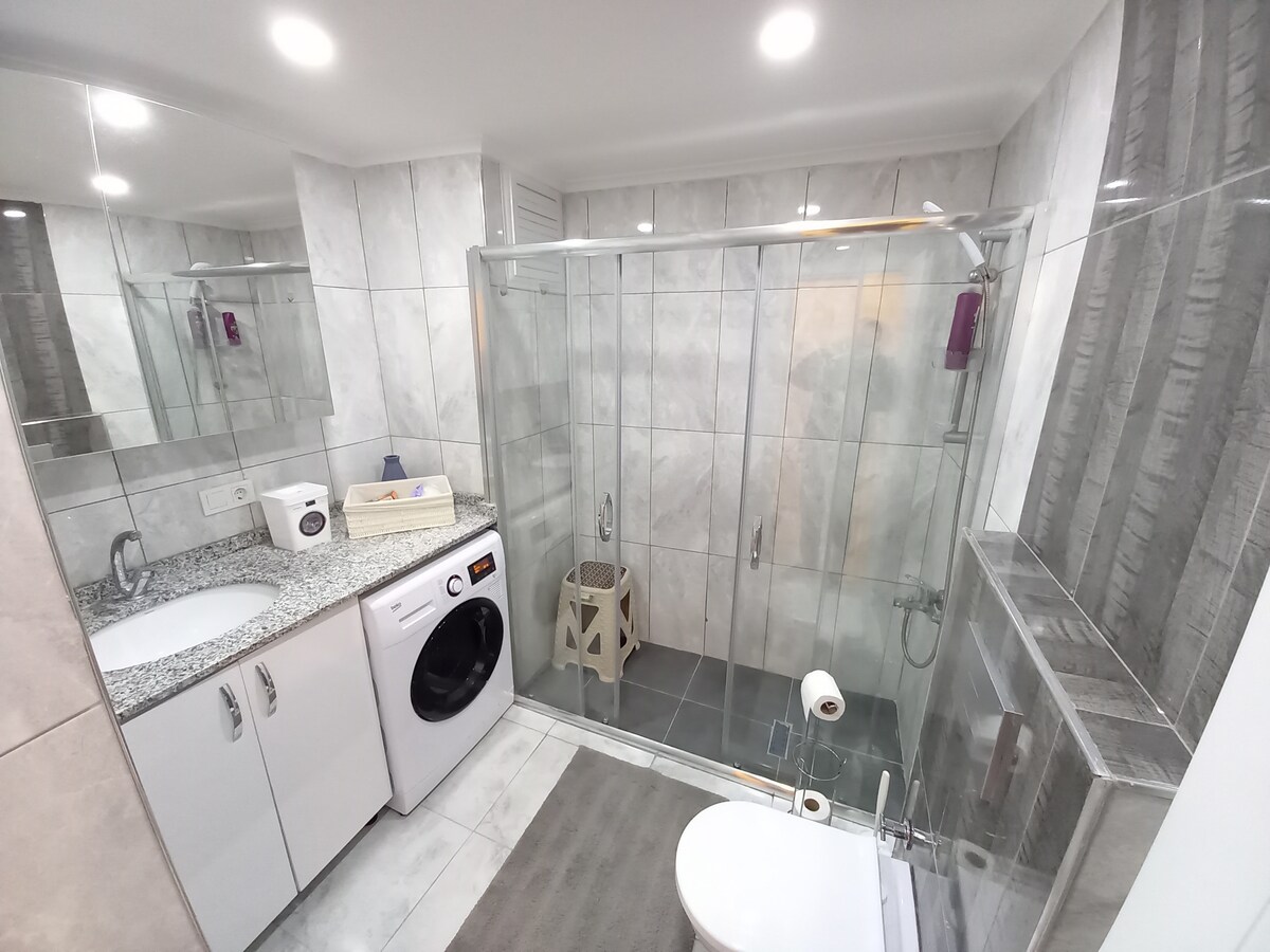 The well-maintained bathroom features a glass shower enclosure with modern fixtures. A spacious countertop made of granite is complemented by a washing machine and neatly organized toiletries. Soft lighting enhances the clean design, while a plush mat adds a touch of comfort underfoot.