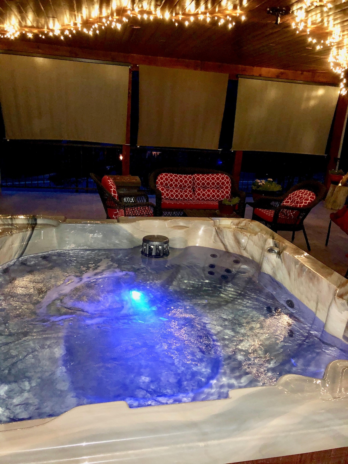 A hot tub is seen with soft blue lighting reflecting on the water's surface. Surrounding the tub are cozy seating areas with patterned cushions under gently glowing string lights, creating a relaxing ambiance. Privacy shades hang partially drawn, enhancing the secluded atmosphere.