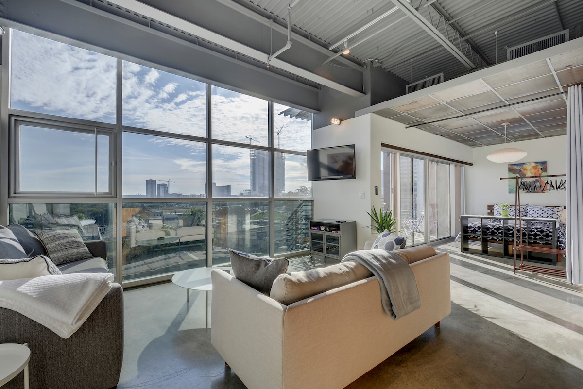 Natural light fills the spacious living area through large windows, offering views of downtown Austin. Two sofas provide comfortable seating, and a flat-screen TV is mounted on the wall. A dining area is visible in the background, adorned with art and a modern light fixture.