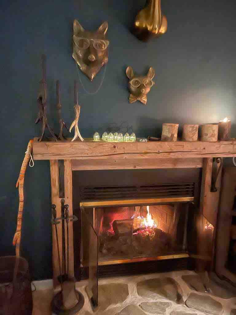 A warmly lit fireplace is positioned within a rustic wooden mantel, surrounded by decorative items including wall-mounted animal head sculptures. The flicker of flames can be seen through a glass front, while the floor features natural stone tiles, enhancing the cozy ambiance.