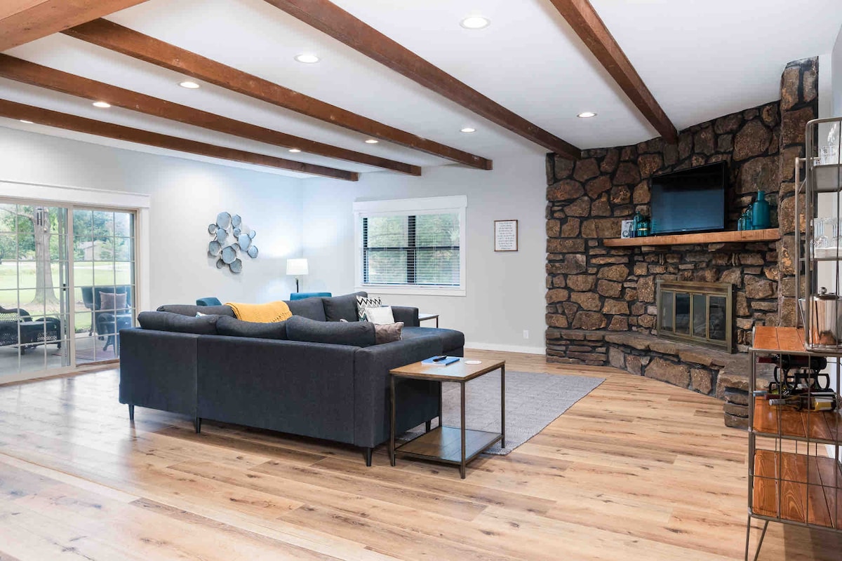 An open living area is highlighted by wooden beams across the ceiling and a stone fireplace. A dark sectional sofa is positioned around a coffee table, while large glass doors lead to the outdoor space. Natural light fills the room, creating a bright atmosphere.