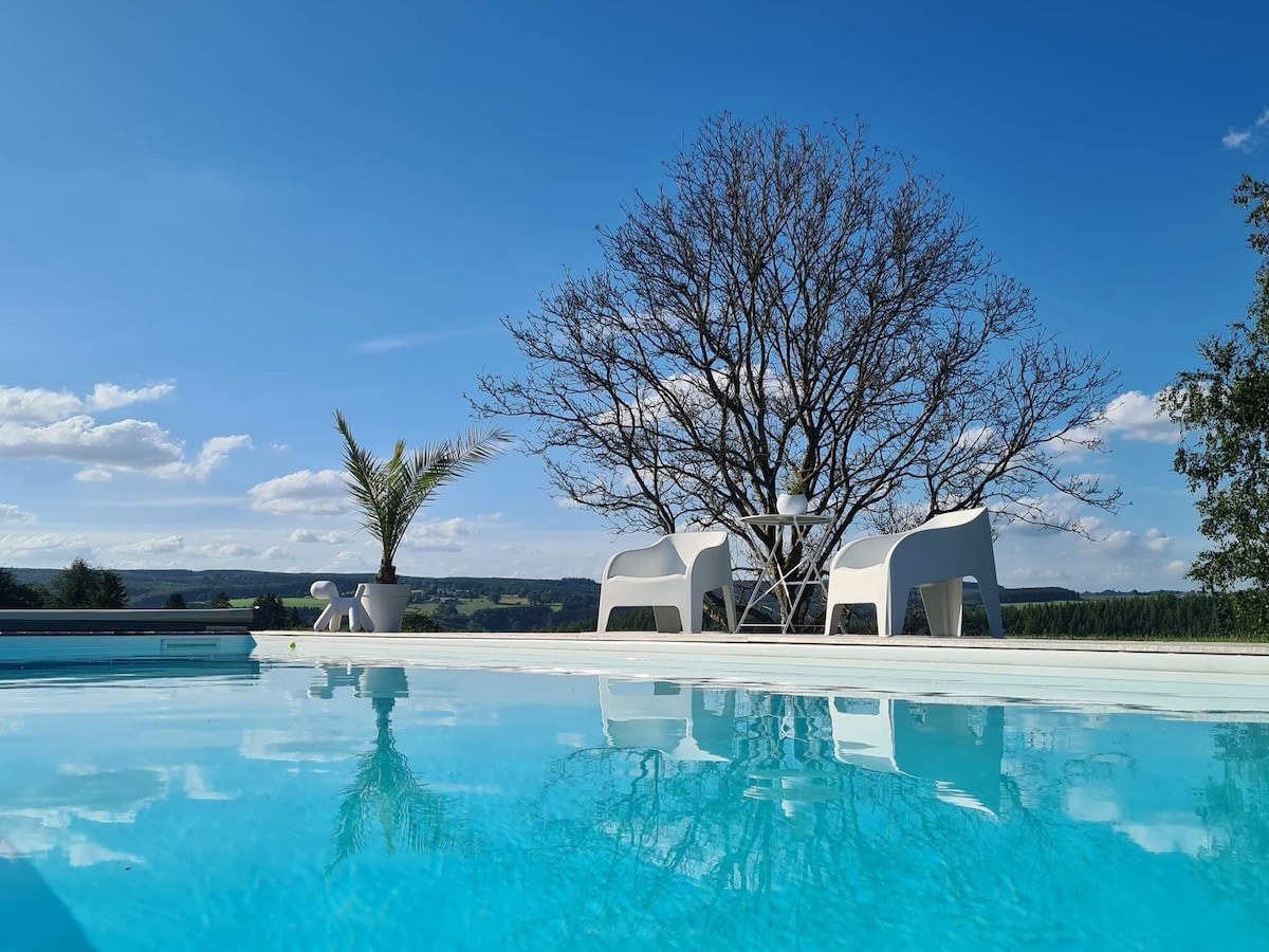 A serene outdoor space is featured, showcasing a pool with clear blue water reflecting the sky. Two white chairs and a small table are positioned near a tree, providing a tranquil spot to relax and enjoy the surrounding landscape.
