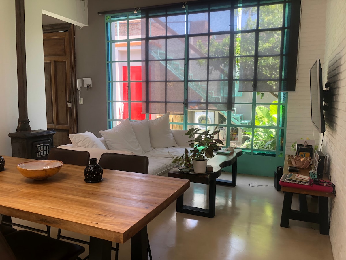 A cozy living area features a comfortable seating arrangement with a light-colored sofa. Large windows allow natural light to fill the space, while a wooden dining table is positioned nearby. A small potted plant adds a touch of greenery to the inviting atmosphere.