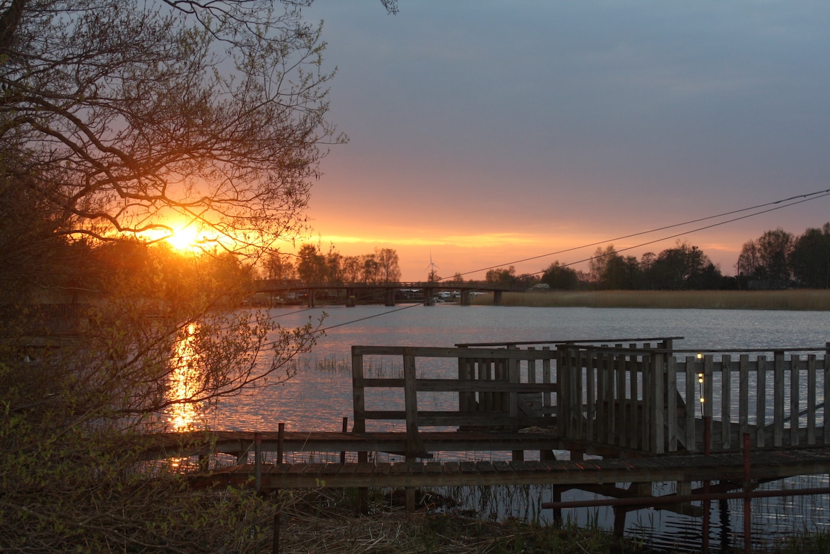 A serene lakeside view captures a vibrant sunset reflecting on the water's surface. Silhouetted trees frame the scene, while a wooden pier extends into the lake, providing a tranquil spot for relaxation. The landscape showcases a harmony of nature and calm water.