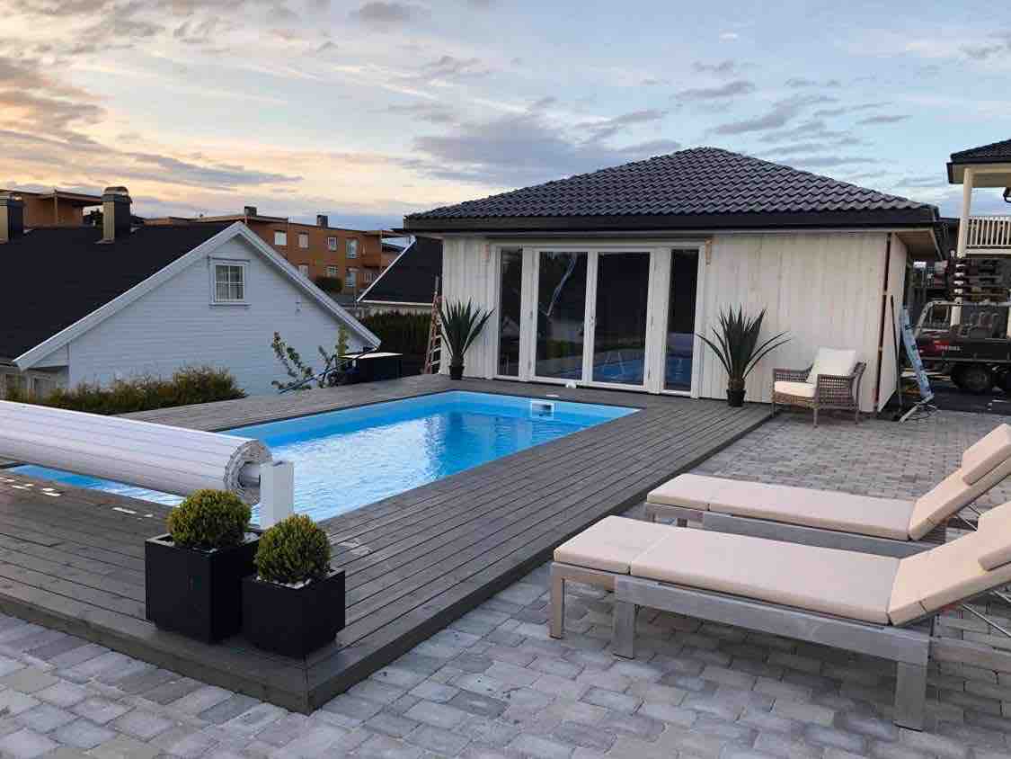 A modern villa features an outdoor area with a heated swimming pool surrounded by decking. Sun loungers are positioned nearby, and the large glass doors of the house provide views into the interior. Green plants add natural elements to the space.